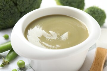 Baby food. Tasty puree in bowl, spoon, peas and broccoli on white table, closeup