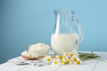 Fresh milk in jug, cottage cheese and chamomile flowers on white wooden table against light blue background. Dairy products