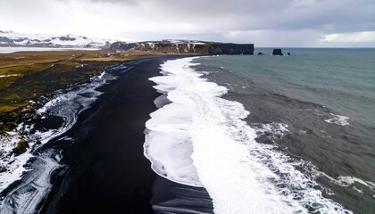 Aerial View Of Black Sand Beach With White Waves Crashing Against The Shoreline And Rocky Outcrops Under A Cloudy Sky