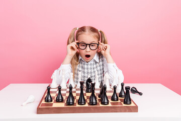 Young schoolgirl in glasses playing chess on a pink background, showing intelligence and curiosity in a fun learning activity