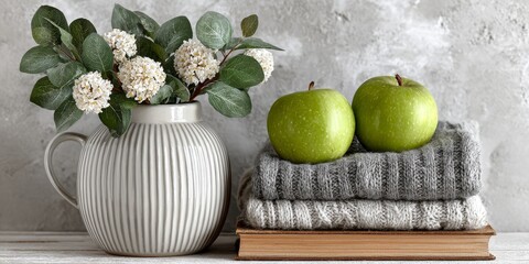 Calm home interior scene featuring green apples on cozy sweaters, a white vase with flowers, and a book against a textured gray wall, evoking seasonal comfort and natural decor