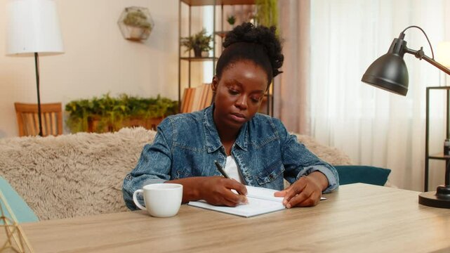 African American young woman sitting at table on home sofa thoughtfully sketches a new logo in notebook, deeply focused on creative design work. Black girl expresses artistic ideas with inspiration.