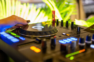 Obraz premium DJ adjusting turntable on professional mixer with glowing buttons, close-up of hand during vibrant party with green plants in background, modern music and nightlife concept.