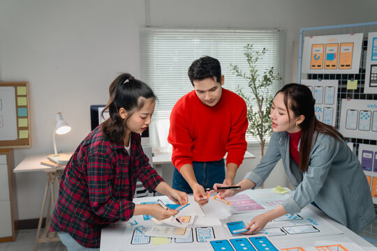 Three Asian UX designers collaborating on user experience and interface optimization for mobile websites and applications, utilizing sketches, wireframes, and prototypes in a bright office