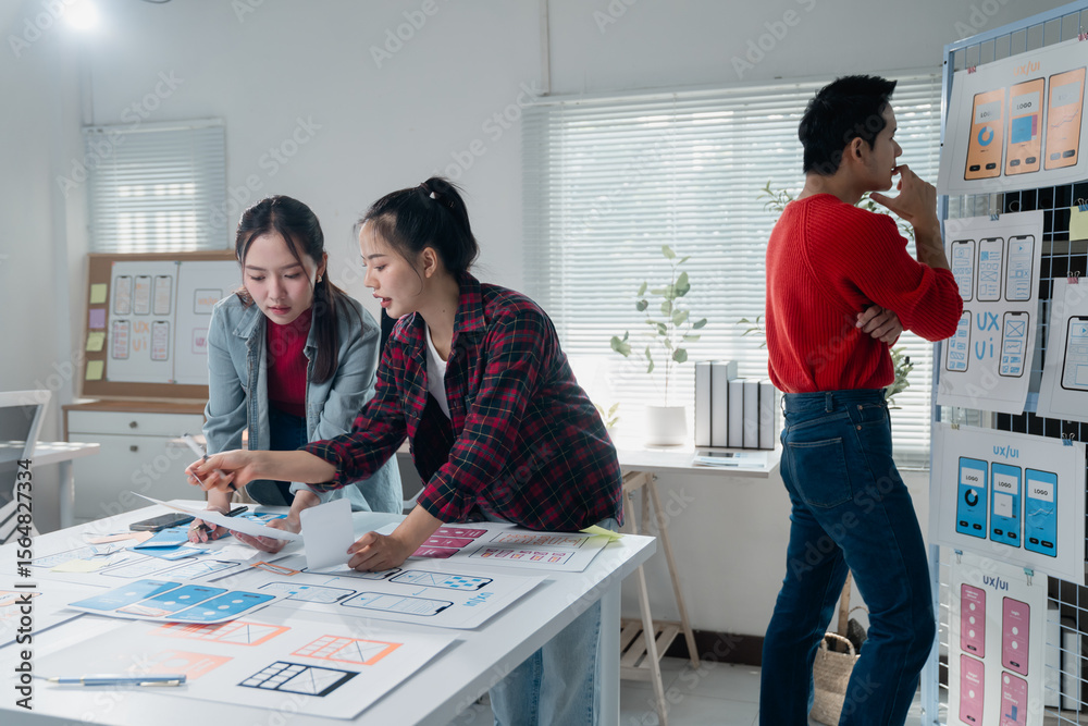 Poster two ux/ui designers analyzing wireframes and prototypes for a new mobile app while a third designer 