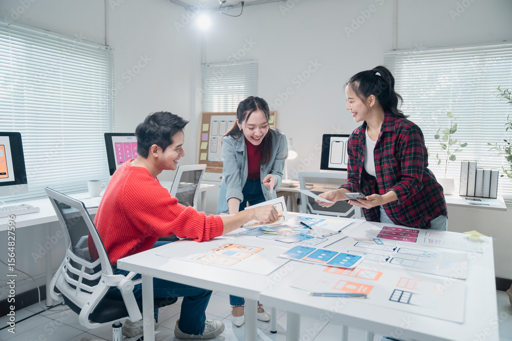 Poster three asian ui/ux developers collaborating on mobile app design in a modern office, using desktop co
