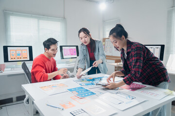 Three Asian UI/UX developers collaborating in a modern office, discussing and comparing printed...