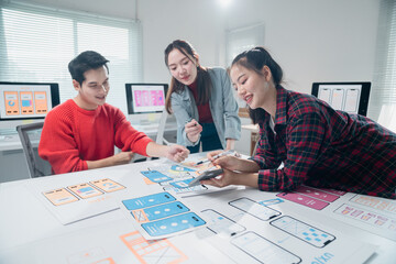 Diverse team of young professionals collaborates on a mobile app design project, using printed layouts and digital tools in a modern office setting, fostering creativity and innovation