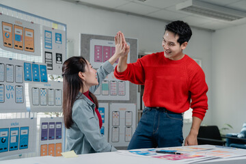 Two smiling Asian colleagues, a male and female UX/UI designer, high-fiving in their office while...