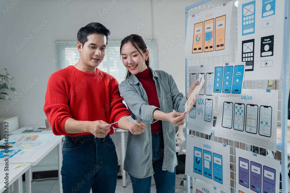 Poster two young asian ui/ux web designers comparing printed mobile app templates at the office, working to