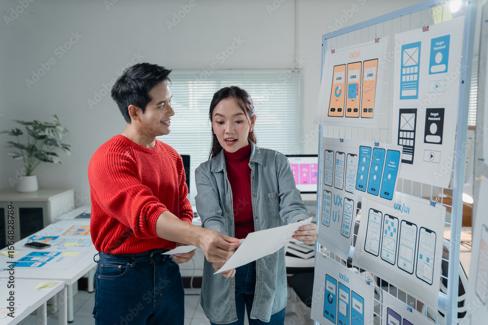 Poster two young asian ui/ux developers reviewing mobile app design wireframes displayed on a whiteboard, d