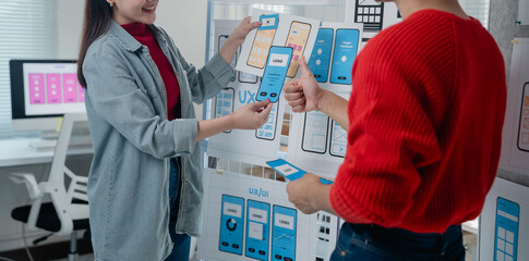 Two Asian ux/ui designers working together developing mobile app design at modern office, analyzing user interface designs hanging on whiteboard, giving thumbs up