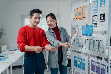 Two young Asian ui/ux web designers comparing printed mobile app templates at the office, working...