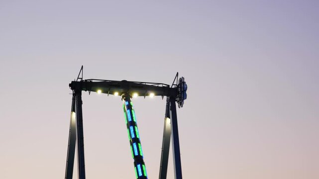 Wide shot of thrill seekers swinging back and forth on a giant Frisbee pendulum ride under a pastel sunset sky