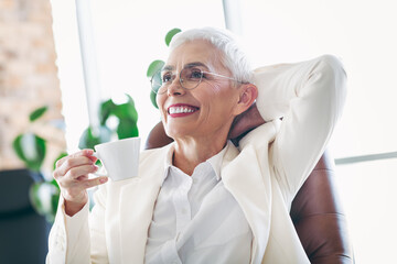 Mature businesswoman in white suit enjoying a cup of coffee in a modern office environment during a...