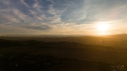 An expansive aerial view of the Casares Valley at sunset, with numerous wind turbines silhouetted on rolling hills, bathed in golden light and distant clouds