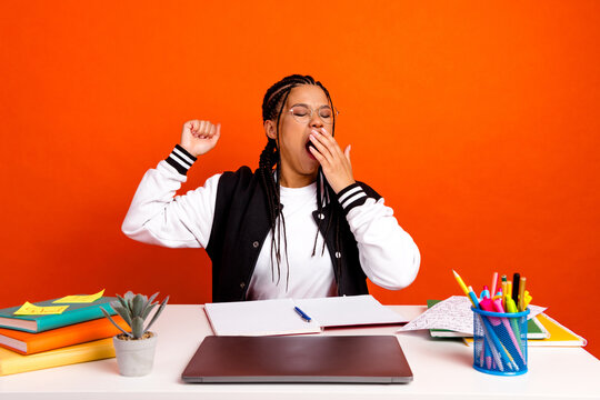 Young college student yawning while studying at desk with vibrant orange background and educational supplies - Powered by Adobe