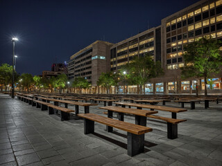 Obraz premium A deserted city plaza with concrete tiles and distant street fixtures faintly lit in the background creating a moody low light space devoid of movement, surface textures and structural