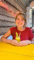 Cheerful Caucasian woman enjoys tropical smoothie under fairy lights, reminiscent of Bali's Galungan festival and exotic retreats