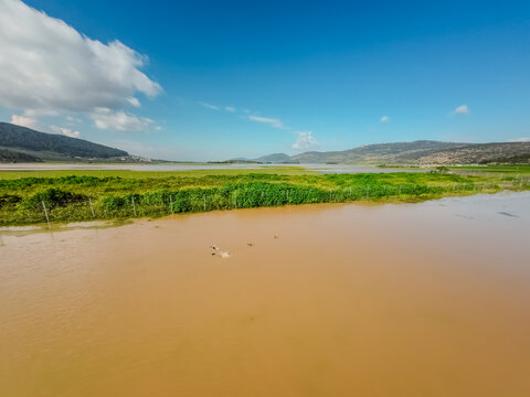 Aerial view of serene waters reflecting the bright sky, bordered by vibrant green vegetation and distant mountains, Biq`at Bet Netofa, Israel.
