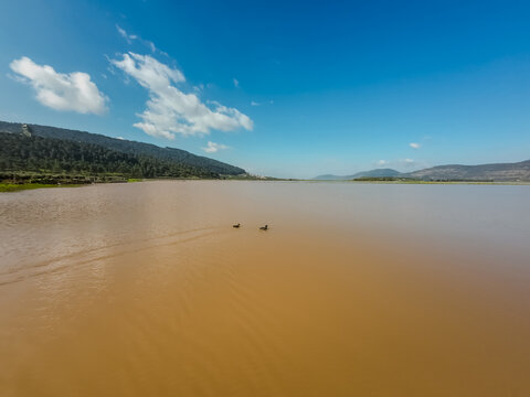 Aerial view of a tranquil, ochre-colored lake reflecting a vibrant blue sky, nestled between rolling hills of deep green foliage, Biq`at Bet Netofa, Israel.