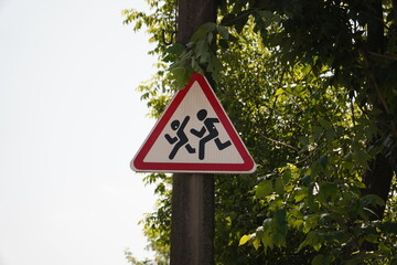 Warning road sign showing children running, mounted on a pole with green trees in the background....