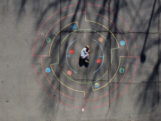 Aerial view of a person curled in the center of a colorful, chalk-drawn target, with concentric circles and vibrant hues contrasting against the grey asphalt, Alessandria, Italy.