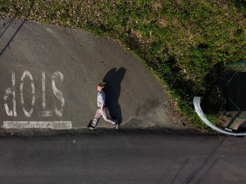 Aerial view of a lone figure casting a long shadow walks past a bold 'STOP' sign painted on the asphalt, contrasting with the adjacent green foliage, Alessandria, Italy.