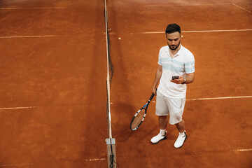 Smartphone in hand, standing, taking a break, bored. Man is playing tennis indoors in the hangar