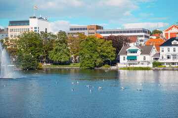Fototapeta premium Scenic view of Byparken park in Stavanger, Norway, with fountain, lake, architecture, and lush summer greenery 노르웨이 스타방에르 Byparken 공원의 분수, 호수, 여름의 풍부한 녹음 풍경