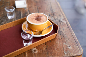 A warm cappuccino in a yellow cup on a rustic wooden table with a glass of water.