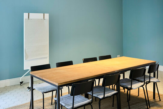 Minimalist meeting room with wooden table, eight chairs, flip chart, and blue walls.