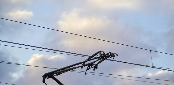 Train pantograph beneath cloudy sky with power lines crossing overhead.