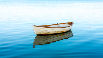 Fototapeta premium A minimalist photo showing a white wooden rowboat motionless on glassy light blue water, reflecting softly under clear, serene sky