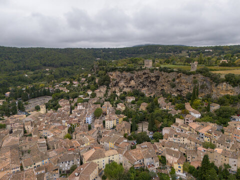 Aerial view of the rustic village nestled at the foot of a dramatic cliff face with ancient towers under a cloudy sky, Cotignac, Provence-Alpes-C&Atilde;&acute;te d'Azur, France.