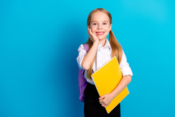 Smiling young schoolgirl with backpack and notebook against a blue background, wearing a white...