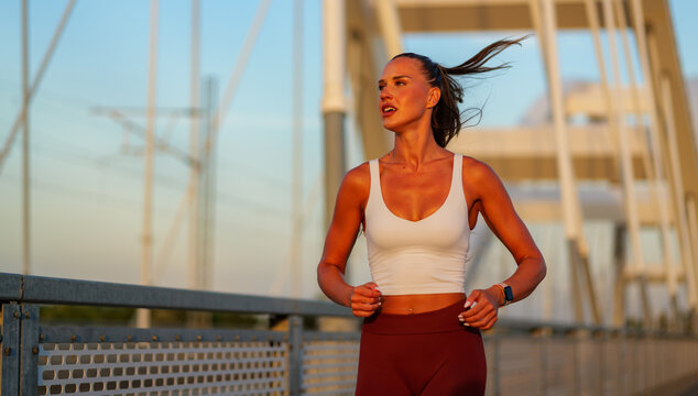 Fit woman jogging across a contemporary bridge during sunset, showcasing determination and fitness. Her hair flows in the wind as she maintains a steady pace, embodying an active lifestyle