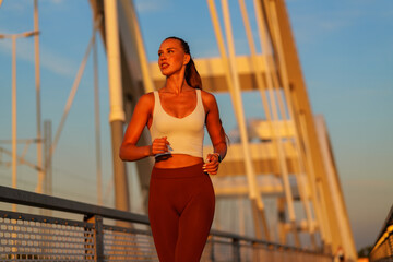 Young sporty woman running on a bridge at sunset, savoring her daily workout routine while...