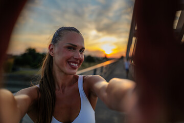 Sporty woman capturing a joyful selfie on a bridge during a stunning sunset after her workout,...