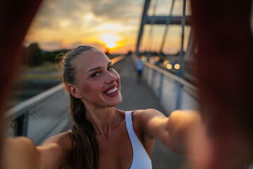 Smiling sportswoman taking a selfie on a bridge during sunset, enjoying the warm golden hour light...