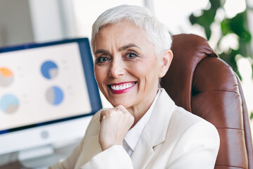 Successful senior businesswoman in professional attire smiling at workplace near a computer screen...