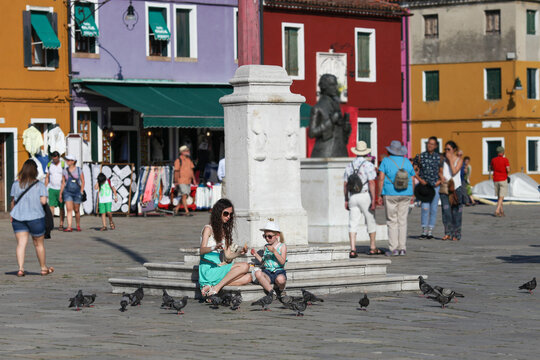 Fototapeta Tourists mother and daughter feeding pigeons at traditional square in Italy, Burano island. Travel with children concept.