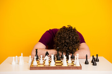 Man resting his head on a chessboard against a vibrant yellow background, embodying frustration and...