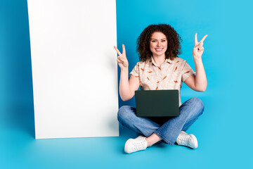 Smiling young woman with laptop sitting on the floor on a vibrant blue background showing peace...