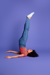 Young woman in casual outfit balancing on her shoulders against a purple background, embracing...