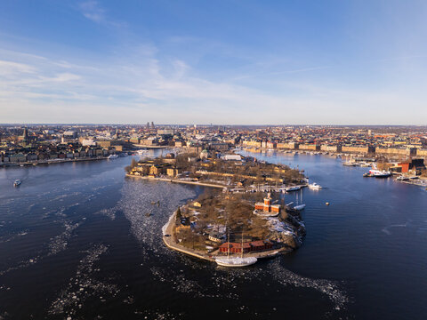 Aerial view of the serene waters surrounding Kastellholmen island, with its historic red buildings contrasting against the icy blue sea, Stockholm, Stockholm County, Sweden.
