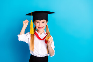 Cheerful schoolgirl celebrating achievement with a medal and graduation cap against a bright blue...