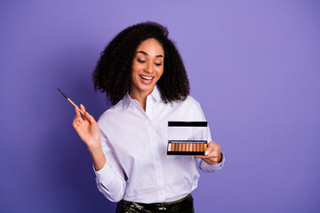 Confident young woman holding eyeshadow palette while gesturing happily over violet background in...