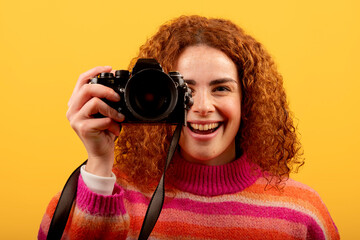Redhead photographer taking pictures with vintage camera smiling on yellow background