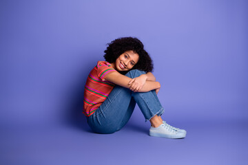 Young cheerful woman sitting against a purple background, wearing casual fashion and expressing joy...